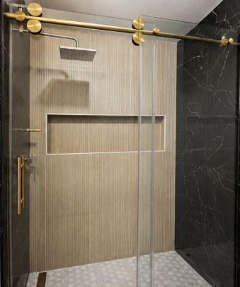 Walk-in shower with gold barn-style sliding glass door, fluted wood-look tile wall and black marble surround