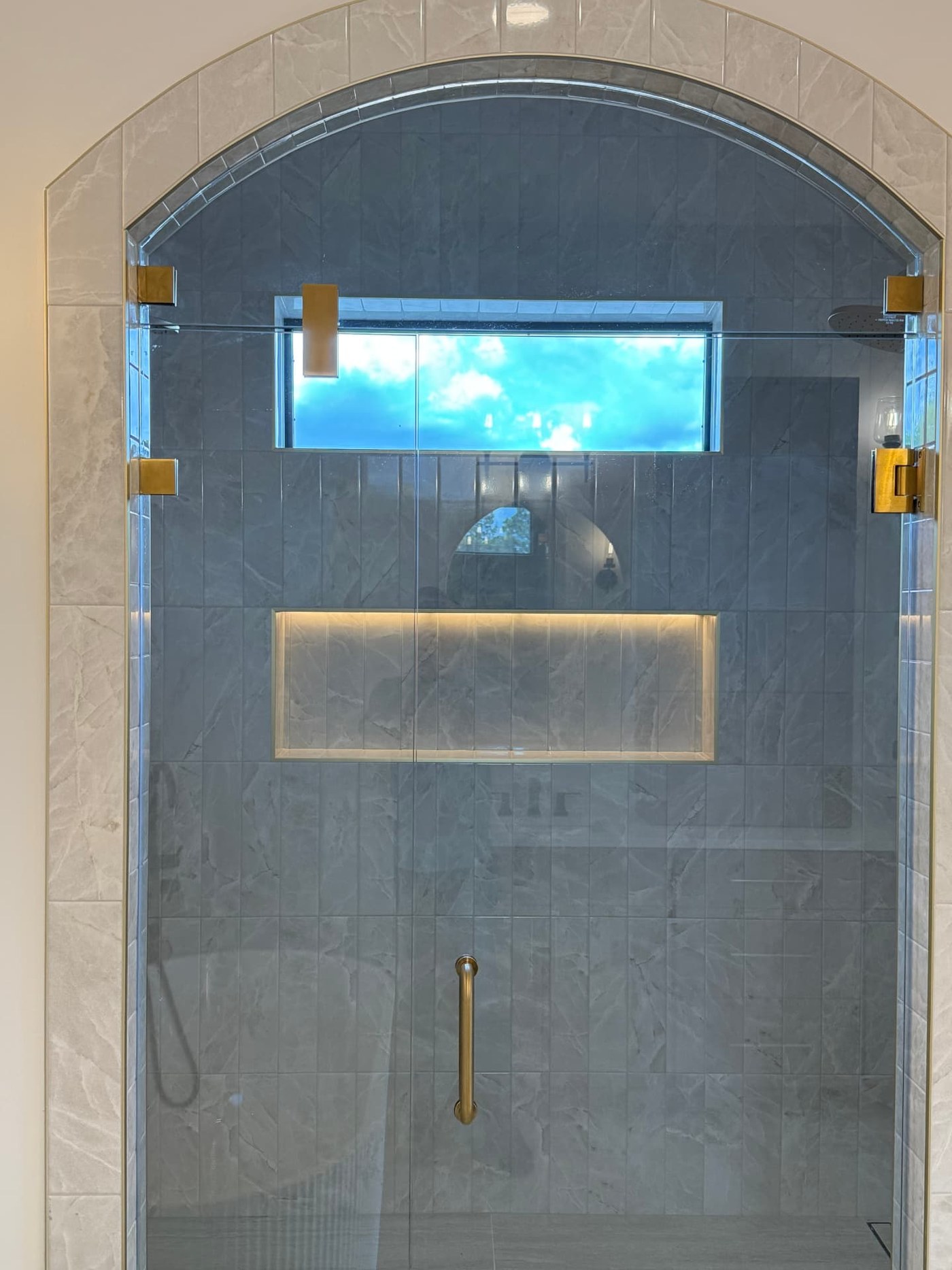 Arched walk-in shower with frameless glass door, brushed gold hardware, vertical stacked stone-look tile and a lit niche