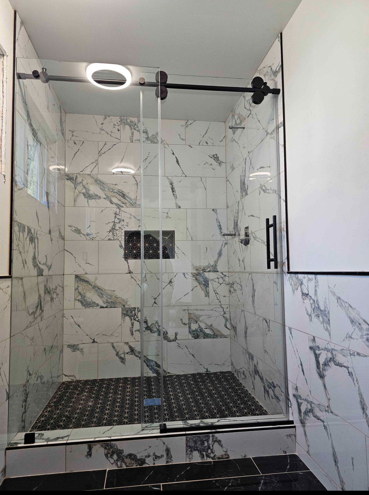 Marble-look shower with a matte black sliding glass door, patterned black mosaic pan and inset niche